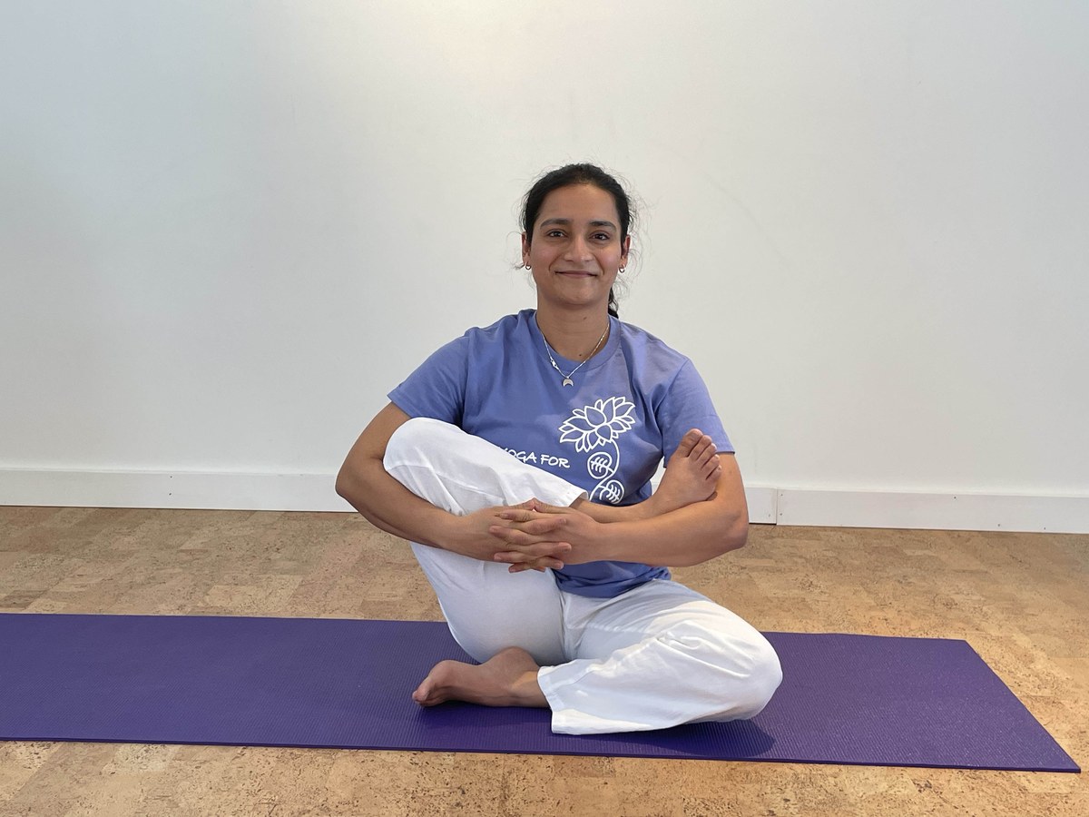 Purnima seated on a purple mat, cradling one leg in a hip-opening stretch